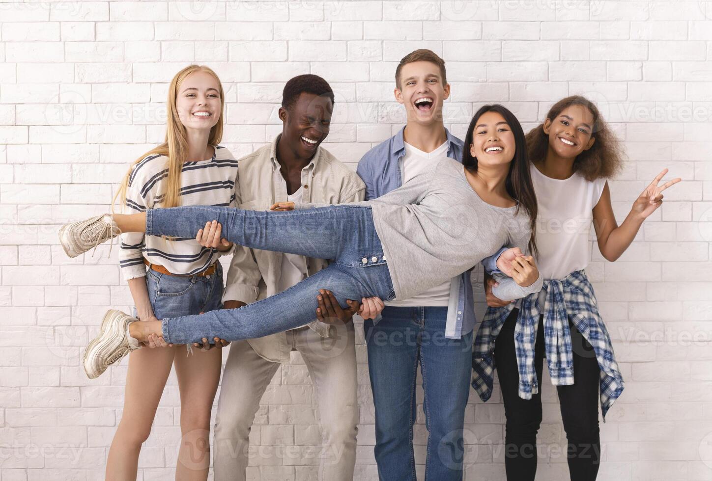Emotional international group of students having fun over white brick wall background, lifting asian girl up photo