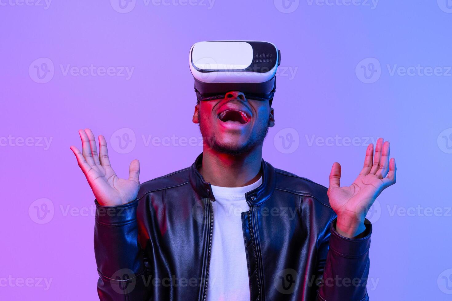 Wow. Amazed Black Guy In VR Glasses Emotionally Reacting To Virtual Reality Experience, Raising Hands And Exclaiming With Excitement While Standing Under Neon Lighting Over Purple Background photo