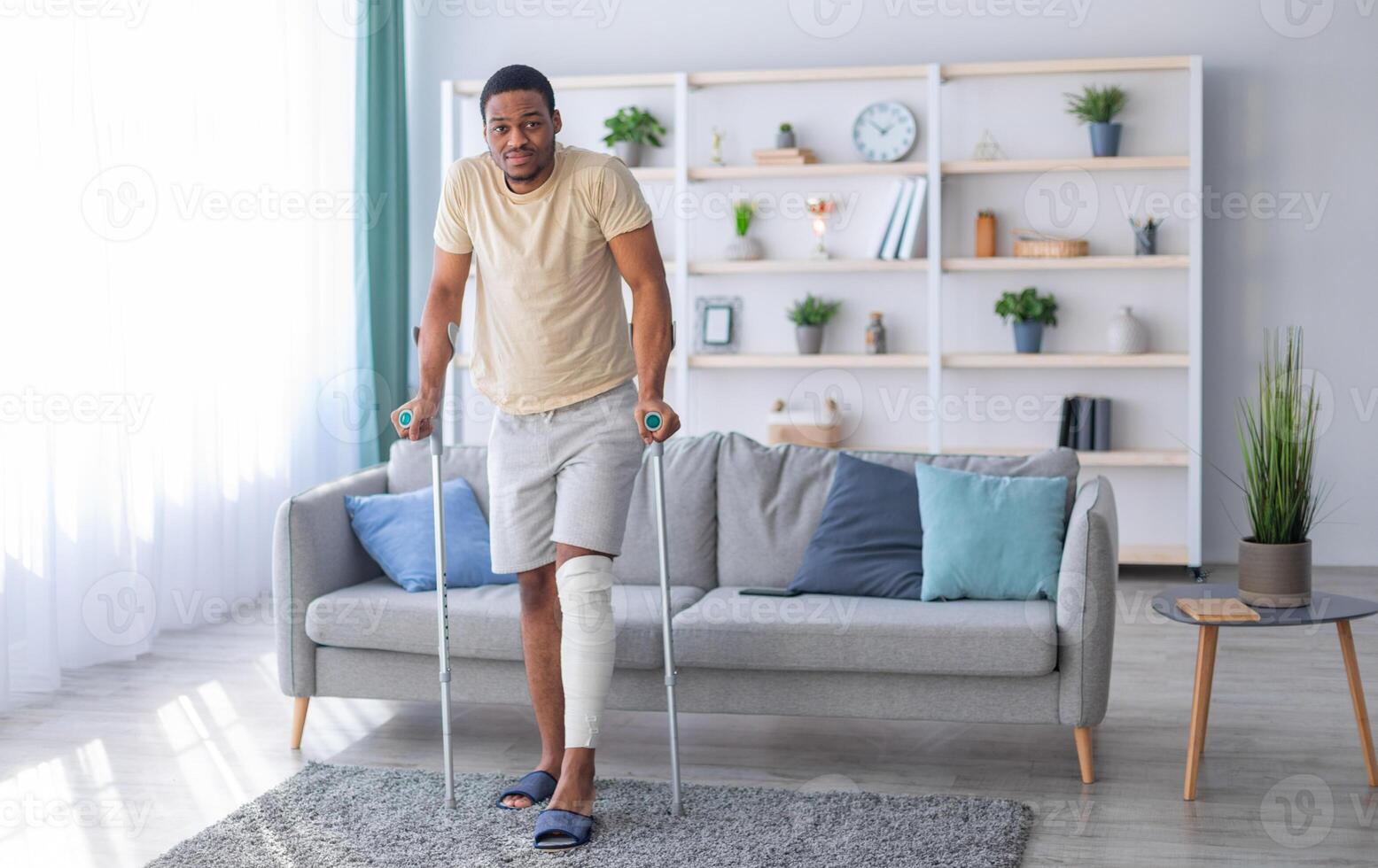 Young black guy with plastered leg walking with crutches, suffering from pain at home, copy space. Full length portrait of millennial Afro man having fractured limb. Health care concept photo