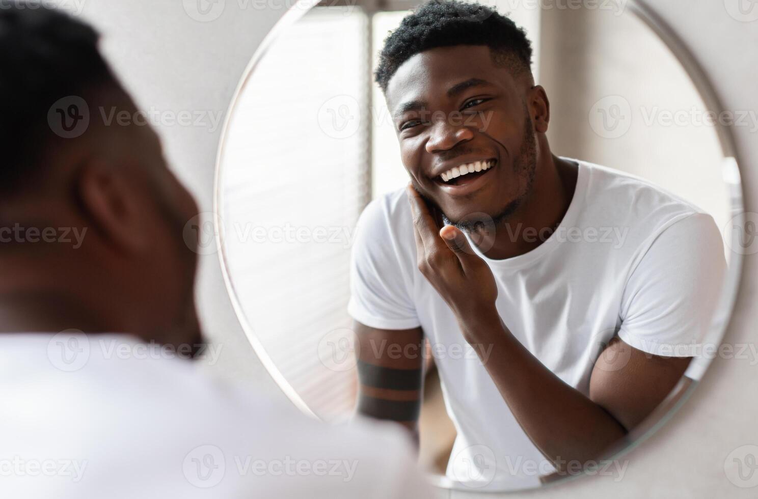 Unshaven African American Guy Touching Face With Bristle On Chin Before Shaving Smiling To His Reflection In Mirror Standing In Modern Bathroom At Home. Male Facial Skincare. Selective Focus photo