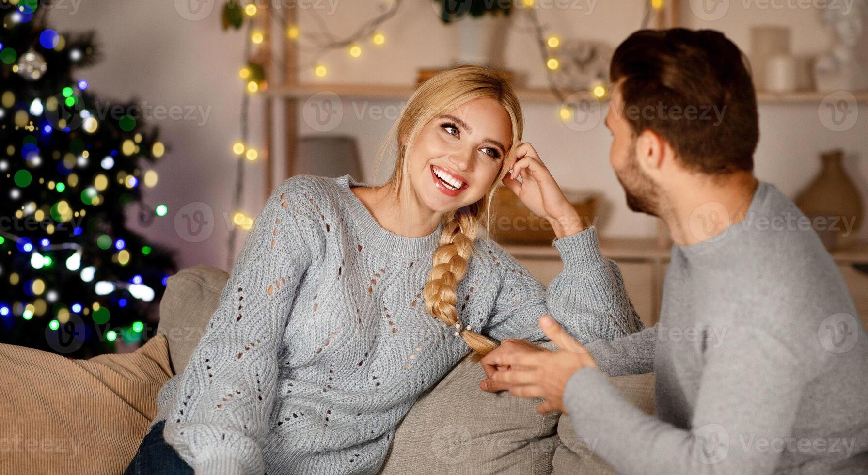 Joyful young man and woman having conversation, laughing and having fun while celebrating Christmas together at home. Happy husband and wife sitting on couch by Christmas tree, talking, copy space photo