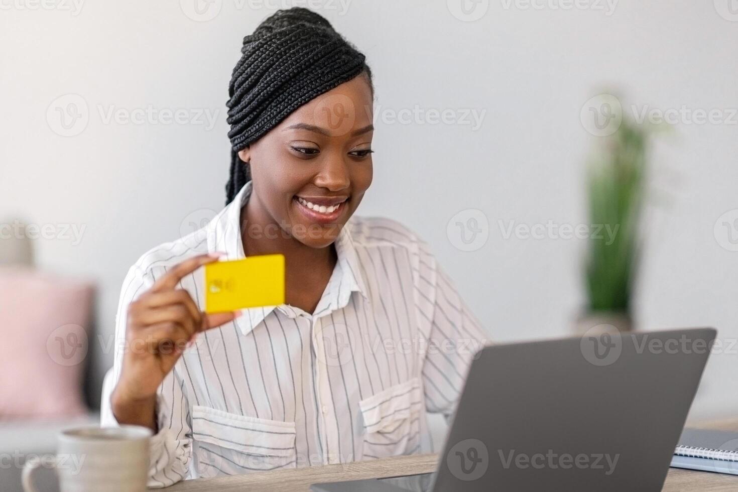 Smiling african american young woman freelancer holding credit card, using laptop, copy space. Happy black lady shopping online, ordering food, cosmetics, clothes from internet while working from home photo