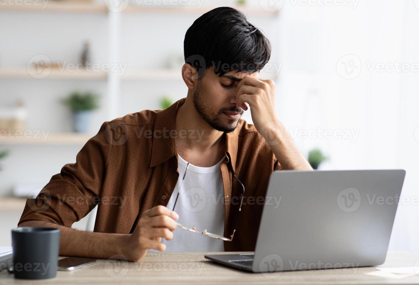 Tired indian man sitting in front of laptop at office, touching his head, suffering from burnout or financial crisis, young guy freelancer having difficulties with job, lack of clients, copy space photo
