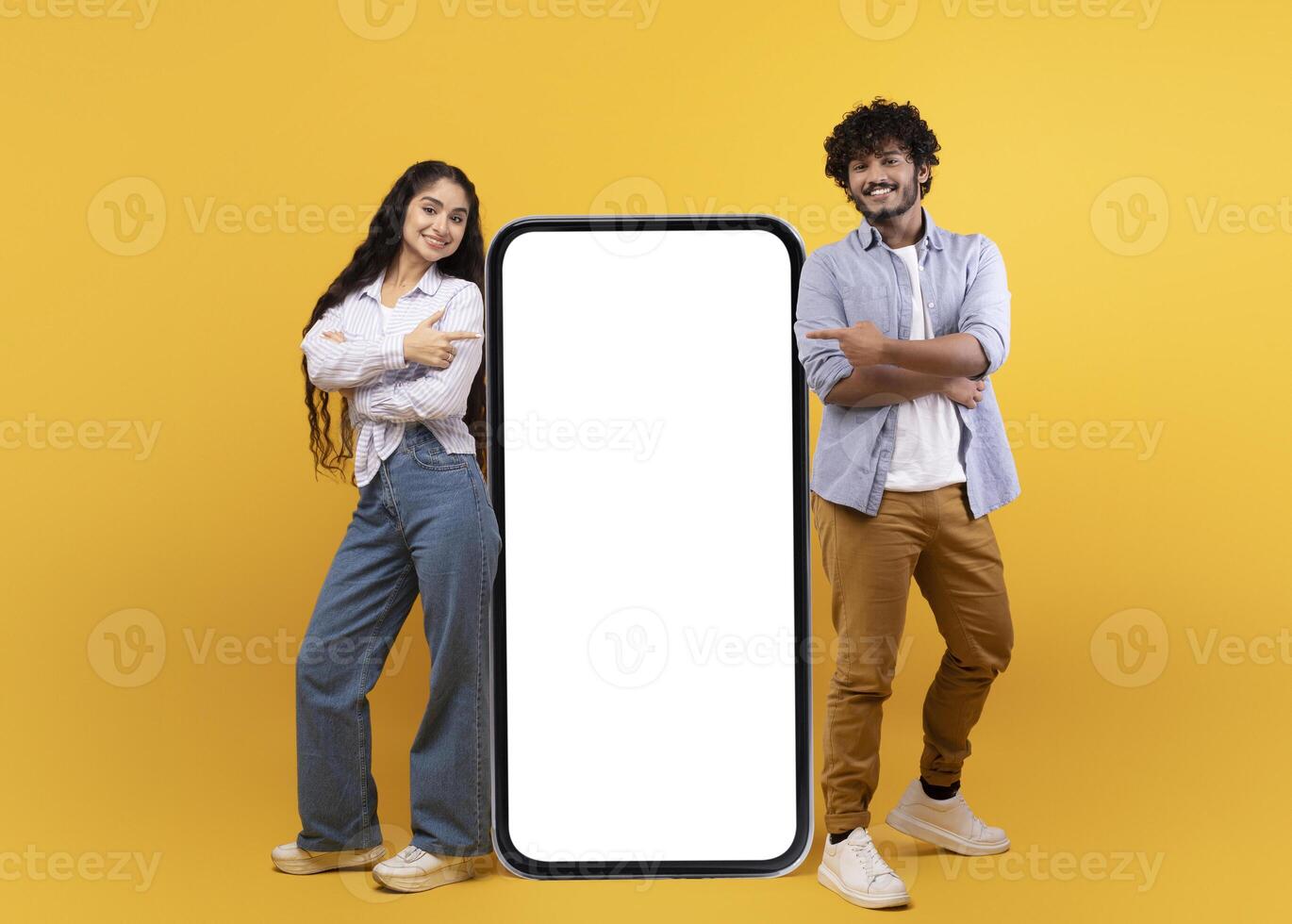 Young indian spouses posing near huge smartphone with mockup, promoting mobile app or website, advertising your service or product over yellow studio background, free space photo