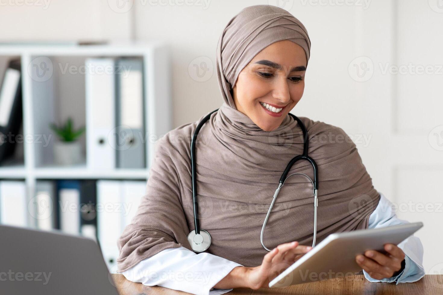 Smiling attractive female doctor woman in hijab using digital tablet in office, sitting at workdesk in front of modern laptop, copy space. Technologies in healthcare industry concept photo