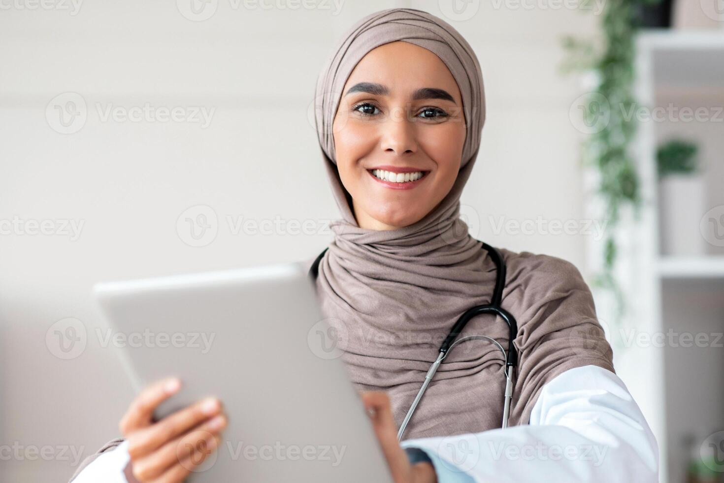 Smiling woman in hijab with stethoscope doctor using digital tablet, sitting at workdesk in clinic, copy space. Female doctor using modern technologies for communication with patients, telemedicine photo