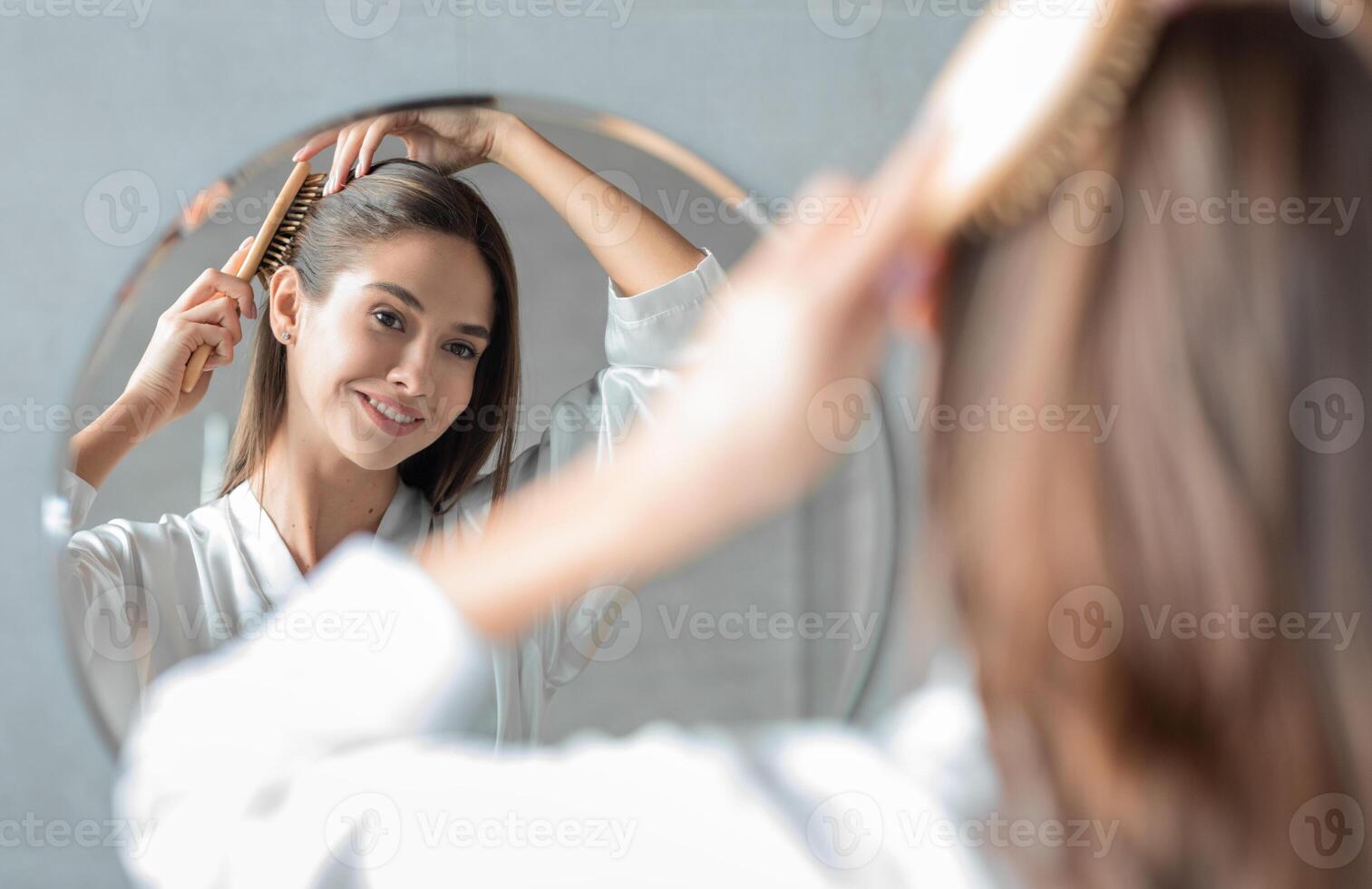 Portrait Of Young Attractive Lady Combing Her Hair With Brush And Looking At Mirror While Standing Near Mirror In Bathroom, Beautiful Female Enjoying Self-Care, Selective Focus On Reflection photo