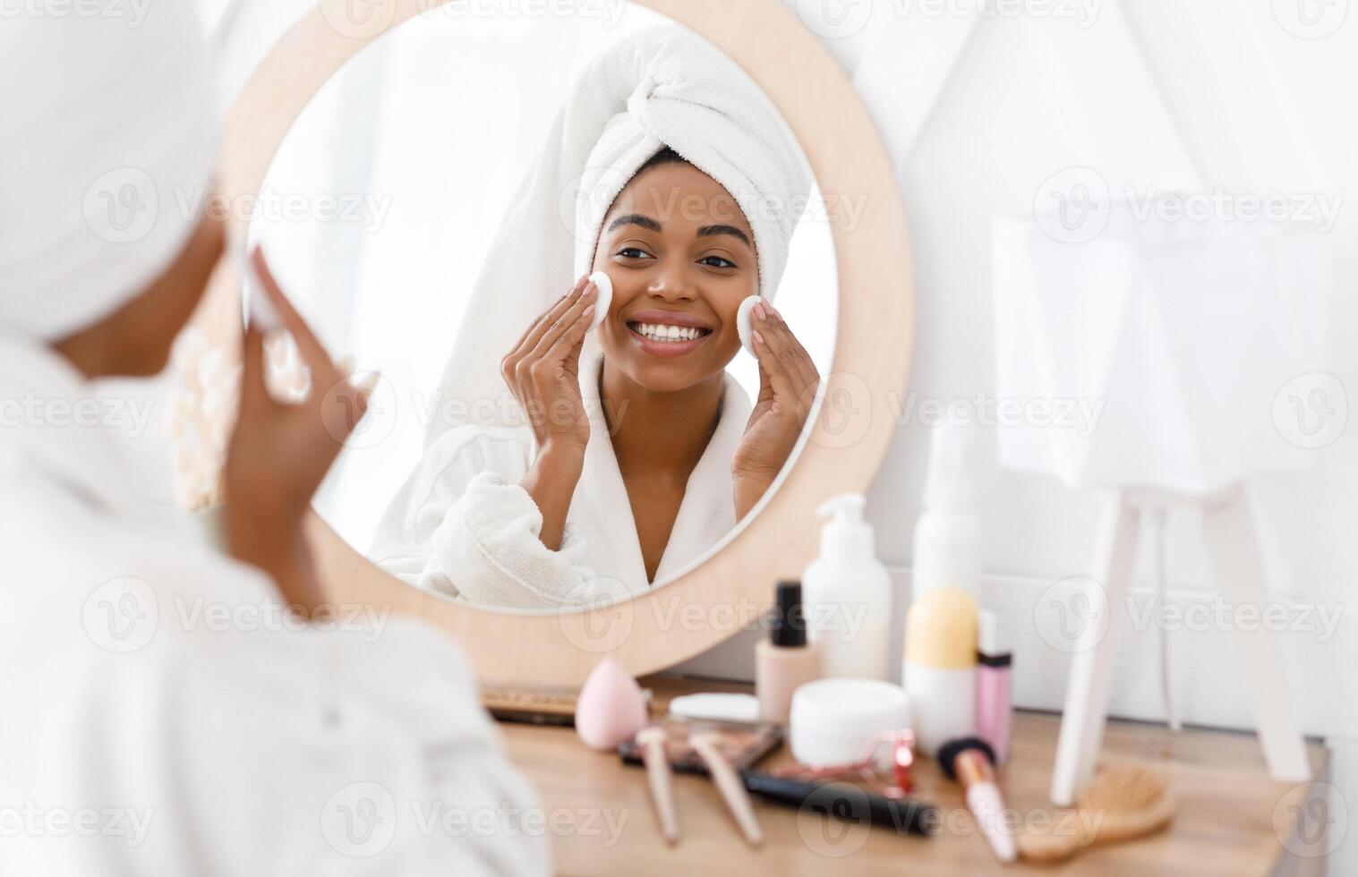 Pretty african american woman removing makeup, cleaning face with cotton pads, looking in mirror while sitting at dressing table photo