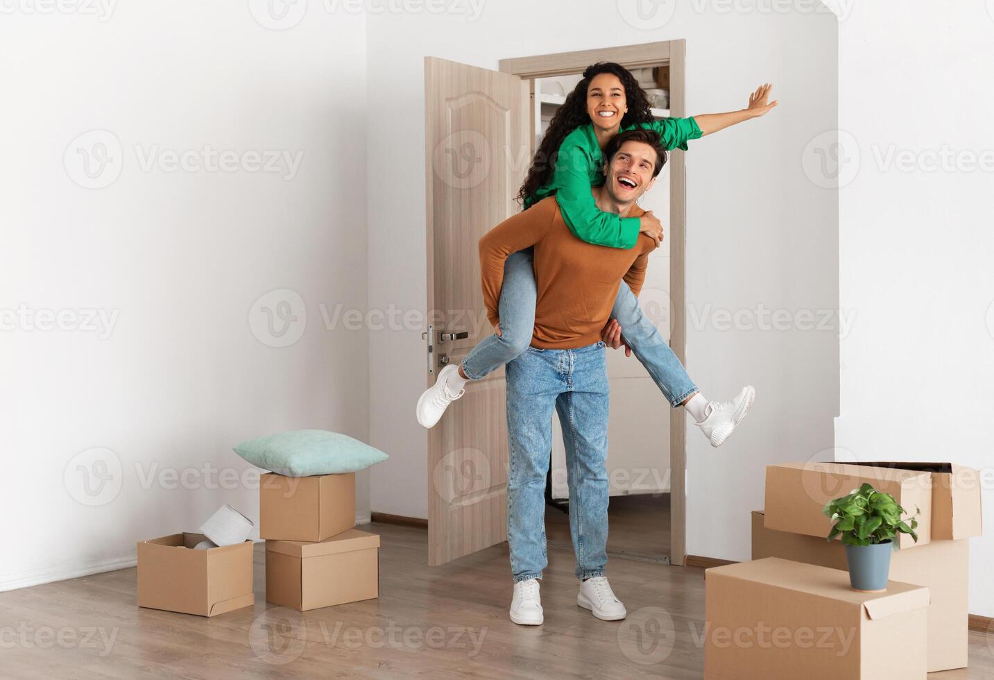 Celebrating Moving Day. Happy couple celebrating relocation to their own home, cheerful guy giving lady piggyback ride, excited young millennial family of two having fun standing in new flat photo