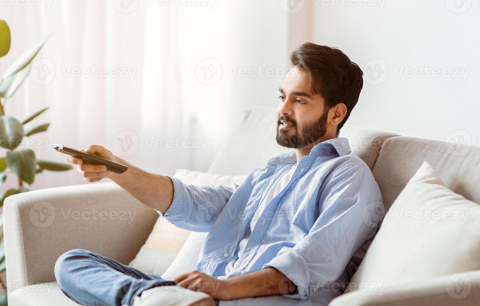 Arab Man Watching TV At Home. Young Eastern Guy With Remote Controller In Hand Resting In Living Room, Sitting On Couch And Switching Television Channels, Millennial Male Enjoying Domestic Rest photo
