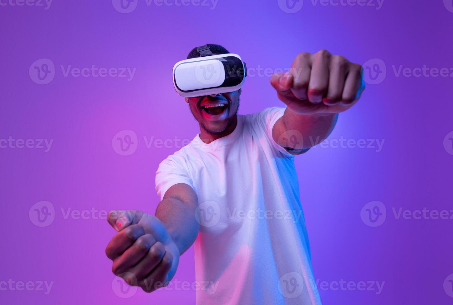 Young african american man playing games in VR-glasses, excited black millennial guy experiencing virtual reality while standing in neon light over purple gradient background, copy space photo