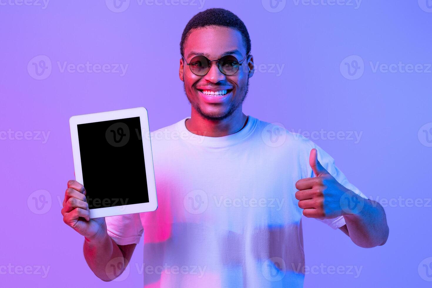 Cheerful Black Millennial Man Holding Digital Tablet With Blank Screen And Showing Thumb Up Gesture, Positive African Guy Recommending New App, Standing In Neon Lighting Over Purple Background, Mockup photo