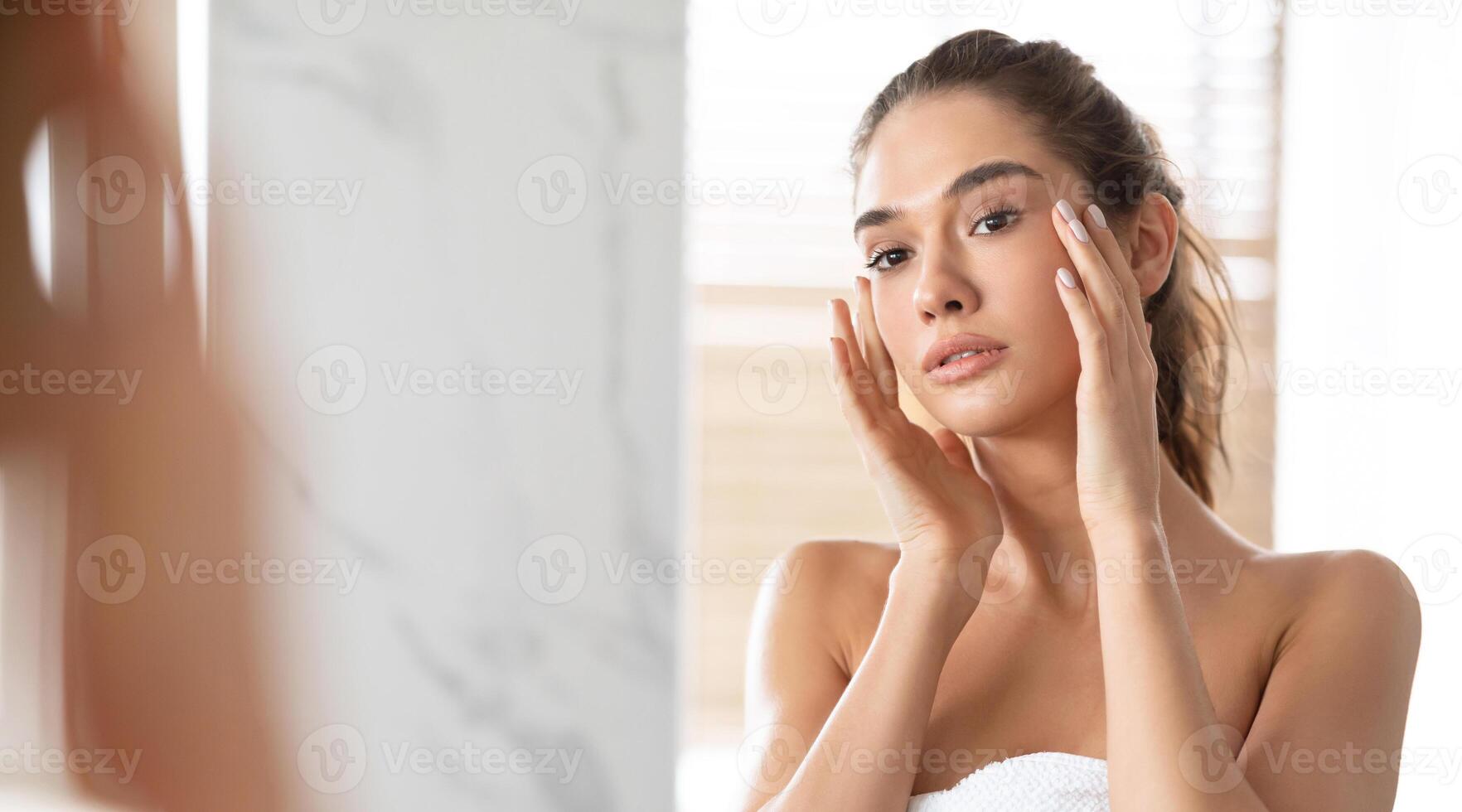 Facial Skincare Concept. Young Woman Touching Face With Perfect Smooth Skin Looking At Mirror Standing Wrapped In Towel In Bathroom At Home. Lady Caring For Skin Indoor. Panorama, Selective Focus photo