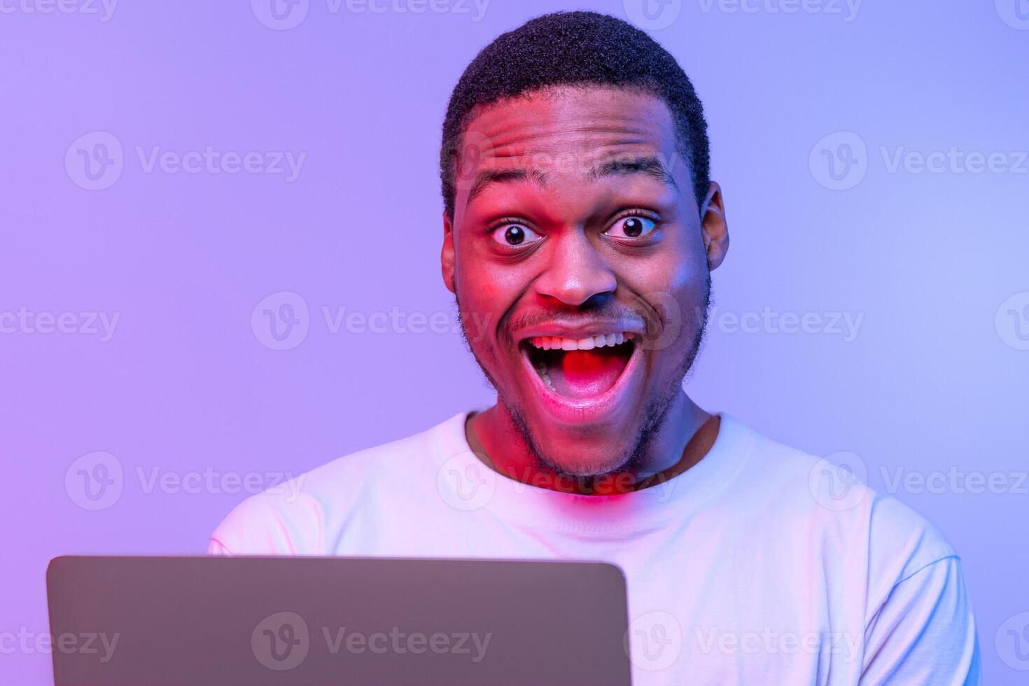 Online Offer. Closeup Portrait Of Of Joyful Young African Man Using Laptop Computer And Looking At Camera With Excitement, Happy Black Guy Standing Under Neon Light Over Purple Background, Free Space photo