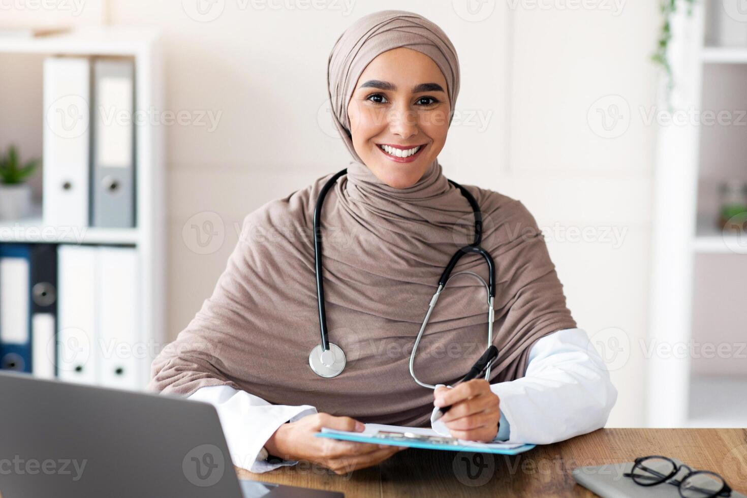 Smiling young woman doctor in hijab taking anamnesis in clinic, sitting in front of laptop, holding medical chart and pen, copy space. Female therapist in head scarf smiling at camera while working photo