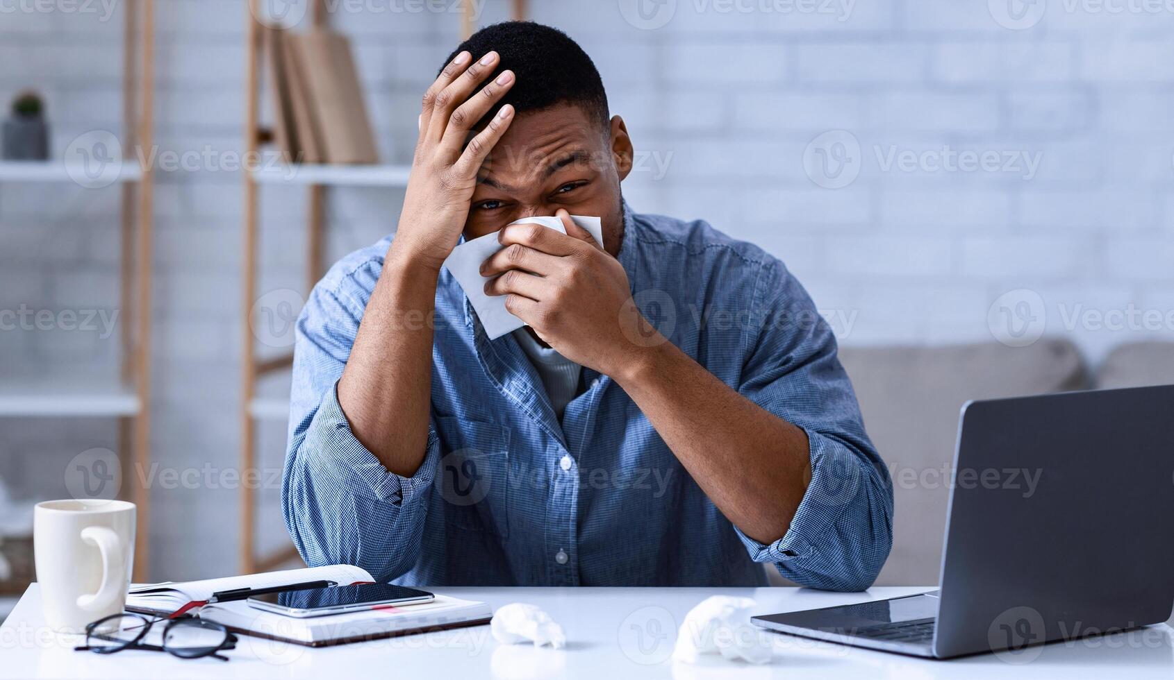 Ill Black Man Blowing Nose In Paper Napkin Sitting At Laptop In Modern Office. Sick Employee At Workplace. Cold And Flu, Rhinitis And Sinusitis Symptoms Concept photo
