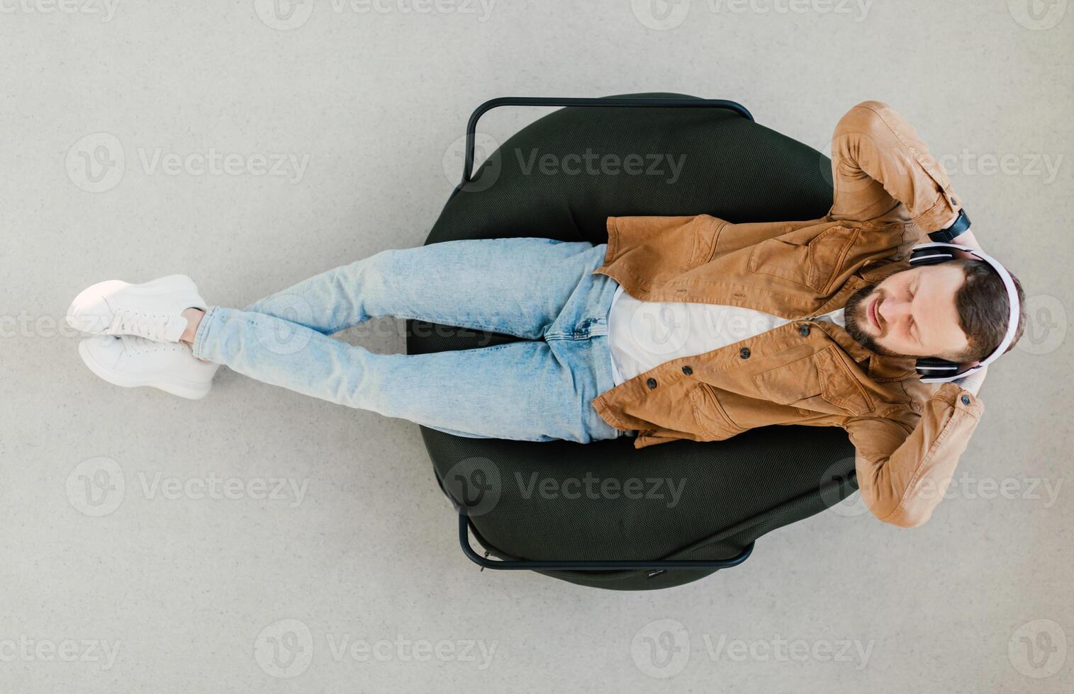 Top View Of Relaxed Guy Wearing Wireless Earphones Listening To Music Or Online Podcast Resting With Eyes Closed Lying In Chair At Home. Weekend Leisure And Relaxation Concept photo