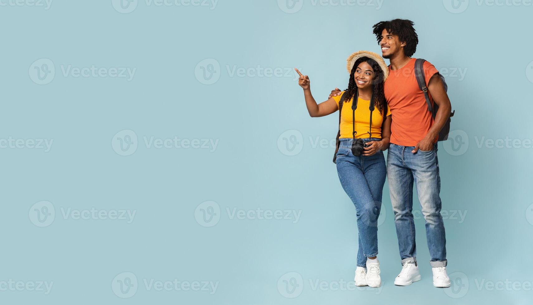 Happy millennial black couple tourists with backpacks and camera embracing and looking at copy space on blue studio background, choosing destination to travel, panorama. Travelling concept photo