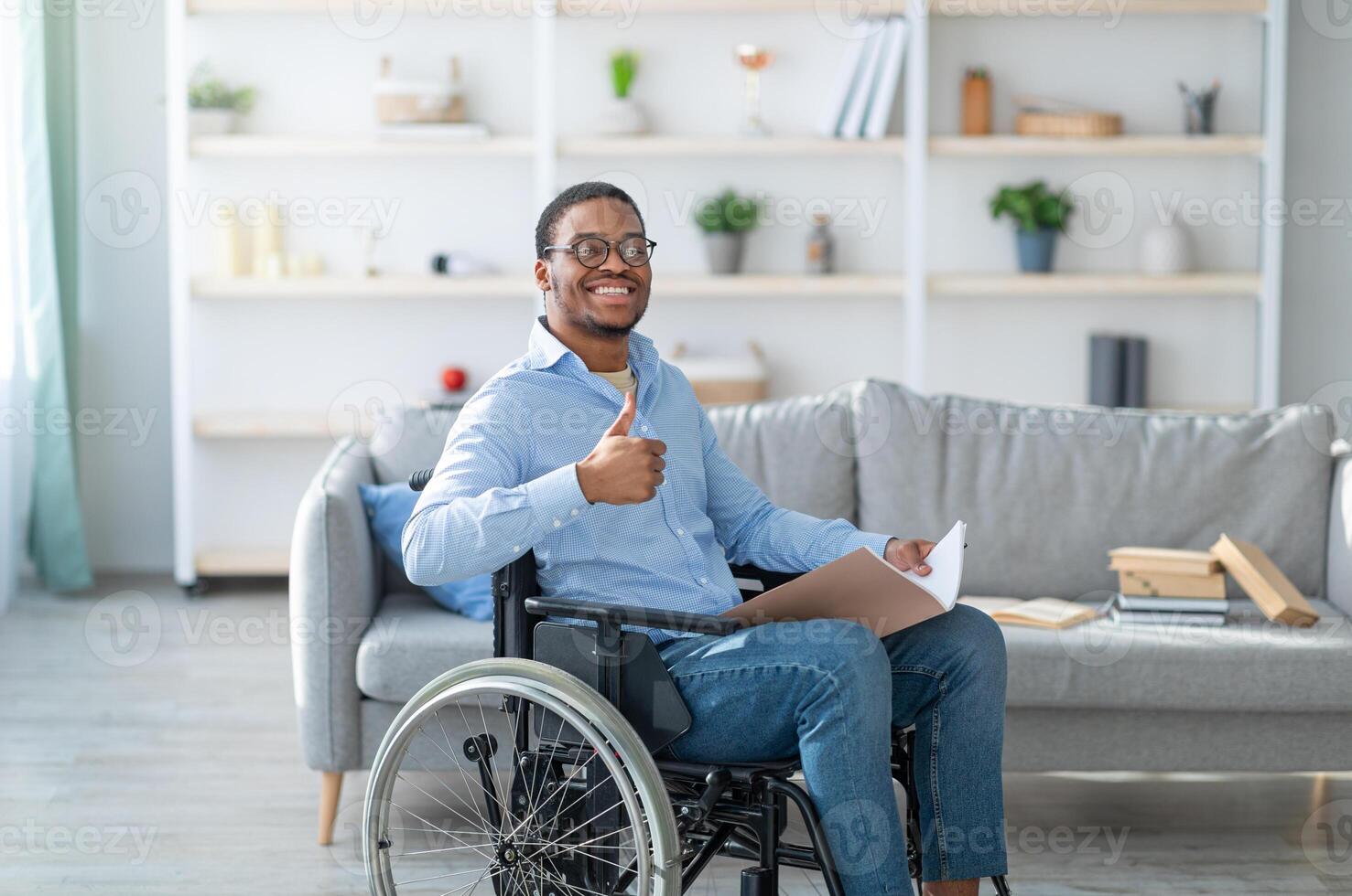 Happy disabled black guy in wheelchair holding notebook, showing thumb up gesture and smiling at home. Millennial African American handicapped guy studying or working indoors photo