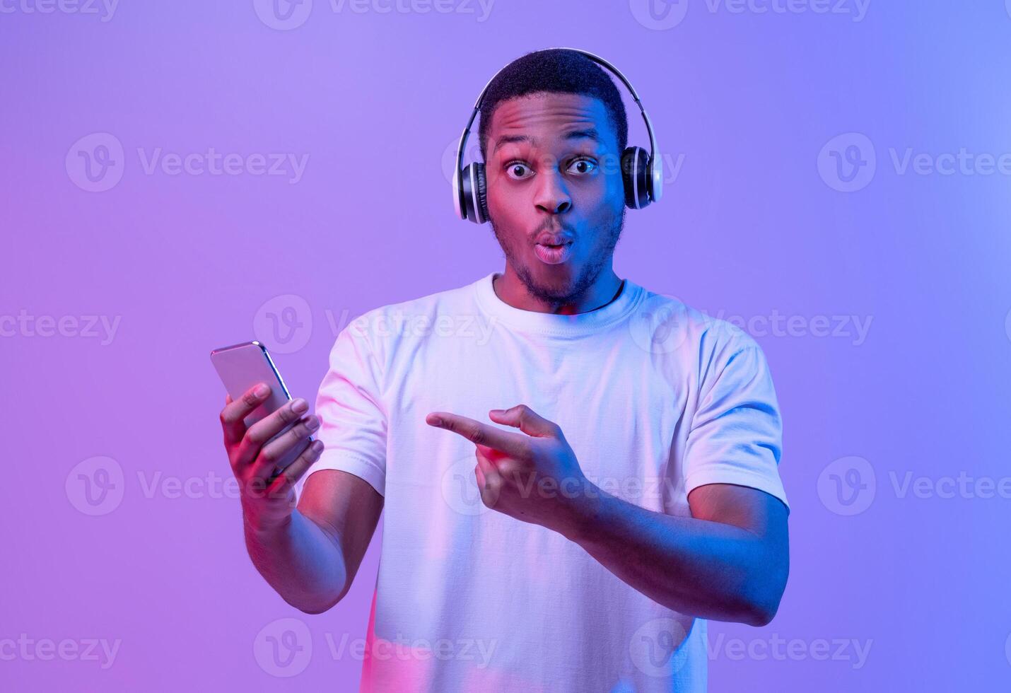 Cool App. Surprised Black Guy In Wireless Headphones Pointing At Smartphone In Hand, Emotionally Reacting To New Mobile Application Or Music Streaming Service, Standing Under Neon Light, Copy Space photo