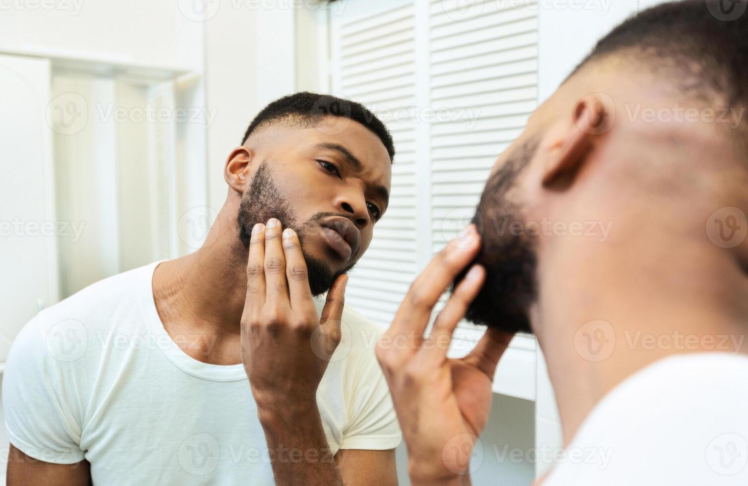 Need to shave. Concentrated african guy looking at his beard at bathroom mirror photo