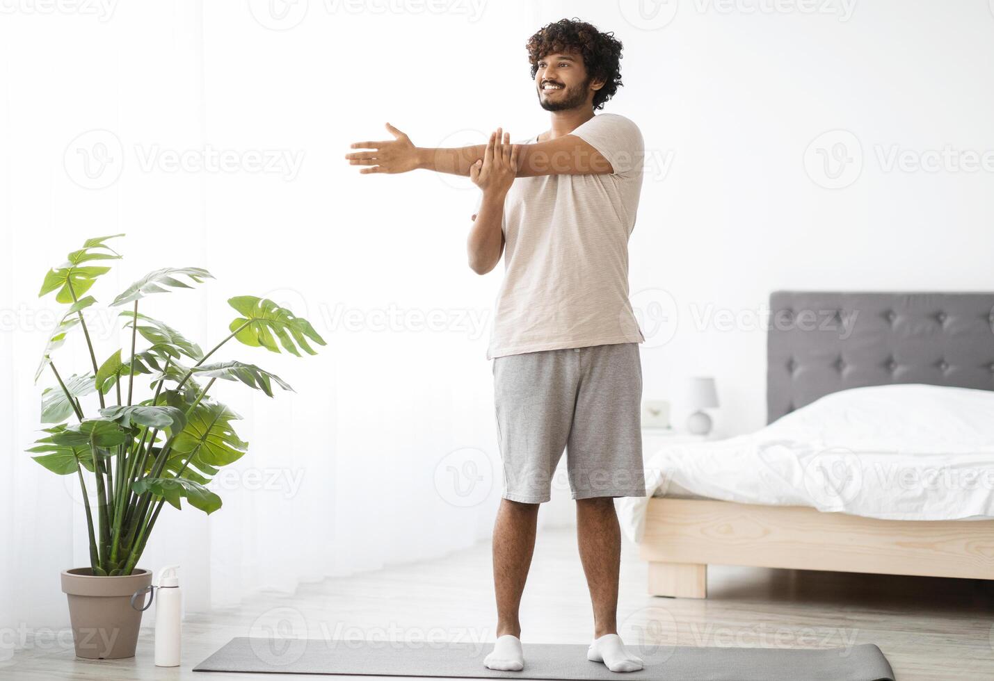 Cheerful millennial indian man in homewear practicing yoga at home, standing on fitness mat and stretching arms, looking at copy space and smiling, enjoying morning workout, panorama photo
