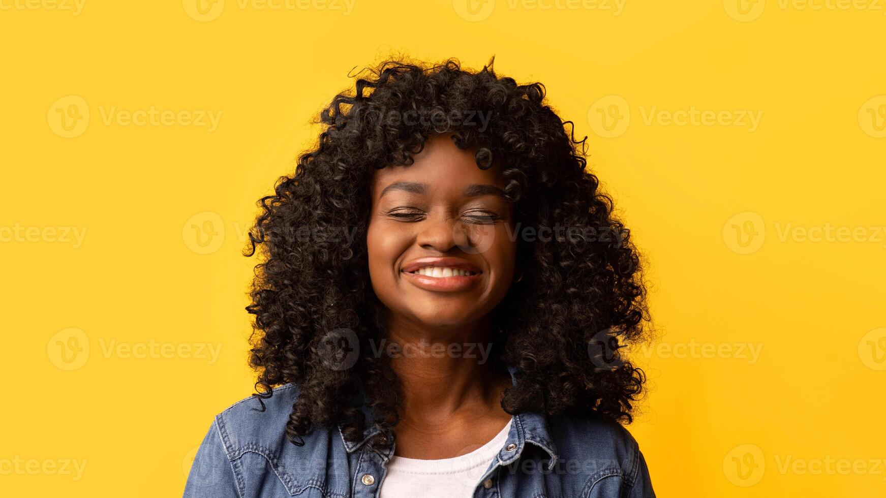 Smiling young african american woman with curly hair on yellow studio background, copy space. Happy beautiful black lady with closed eyes, empty space for advertisement, youth and beauty concept photo