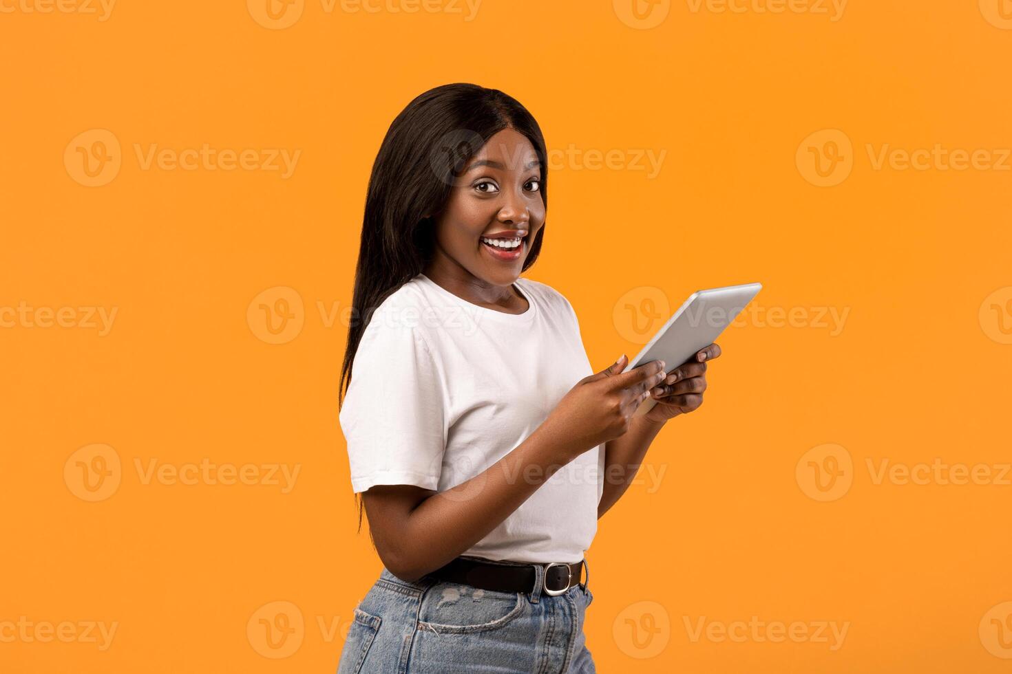 Positive young black woman with modern digital tablet posing on orange studio background, copy space. Cheerful african lady using mobile application on pad, technologies and entertainment concept photo