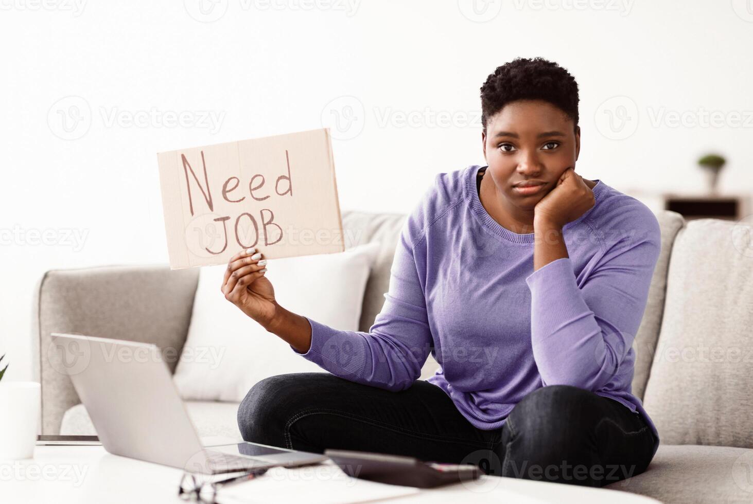 Portrait of unhappy unemployed black woman holding cardboard sign with Need Job written text. Frustrated young lady begging for a work and employment during coronacrisis sitting on couch at home photo