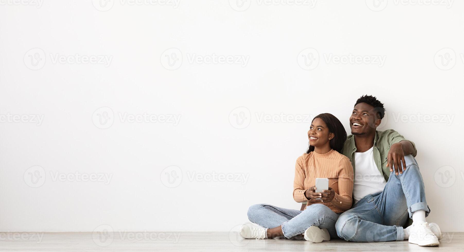 Cheerful millennial black couple with smartphone looking at copy space, sitting on floor by white wall at empty apartment, buying furniture for house online, using mobile app, panorama photo