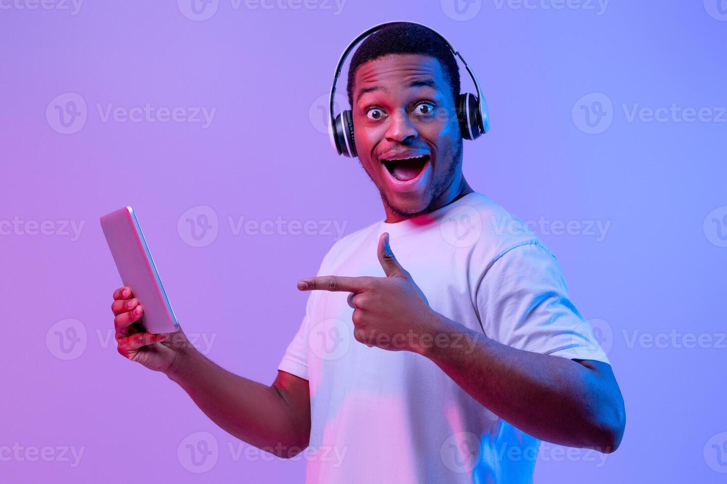 Modern Gadgets For Entertainment. Happy black man in wireless headphones using digital tablet, excited african american guy pointing at device, standing under neon light on purple studio background photo