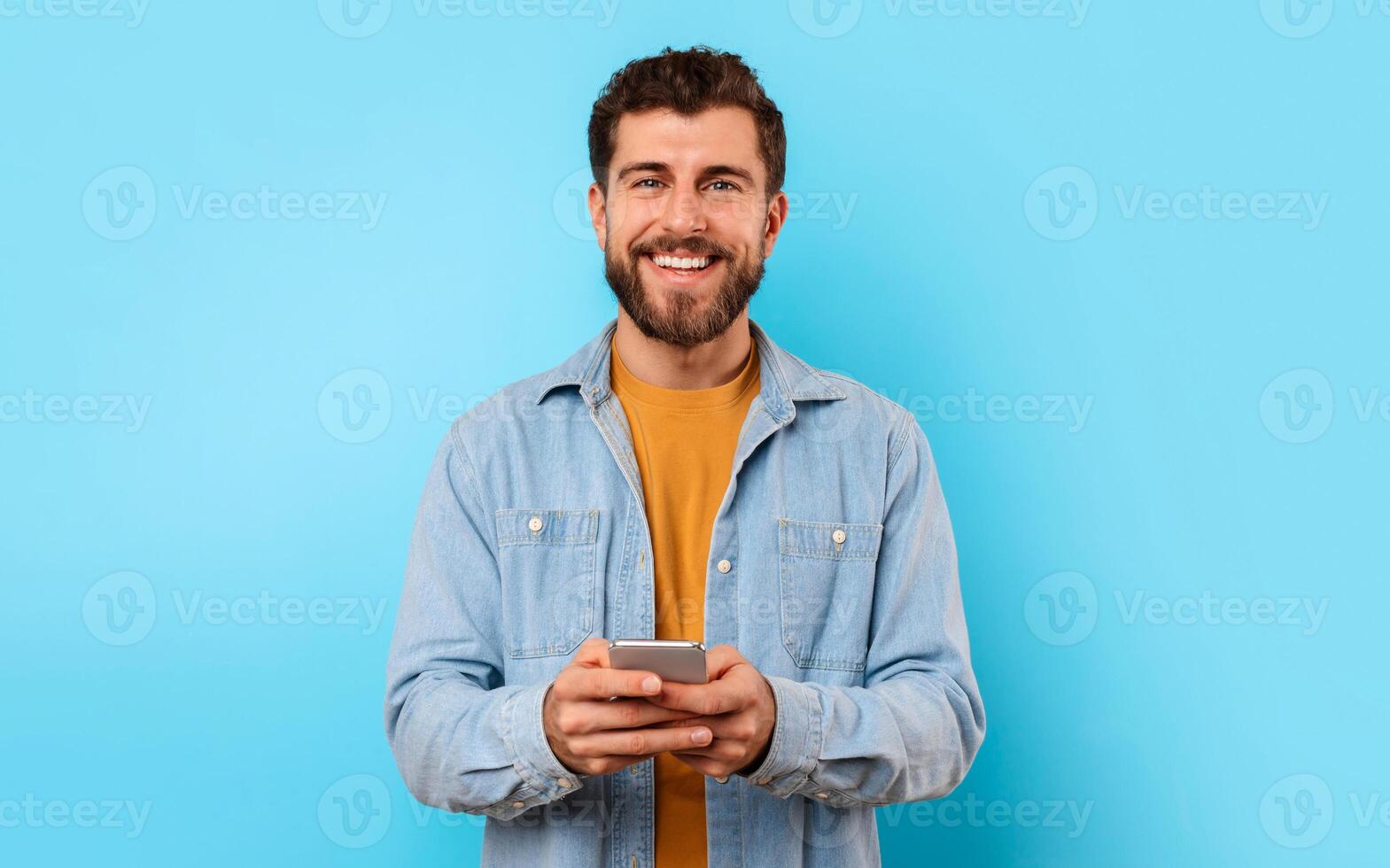 Cheerful guy with smartphone looking at camera and smiling, texting in social media over blue studio background. Positive young man in casual chatting on phone, using new mobile application photo