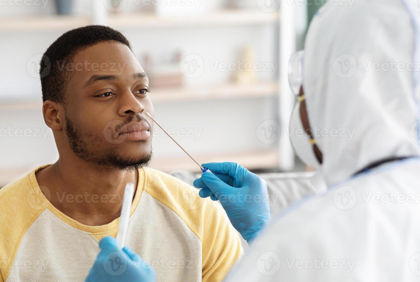 Black guy receiving nasal PCR test while staying home during COVID-19 pandemic, closeup. Unrecognizable doctor in protective suit taking nasal swab for young african american man patient photo