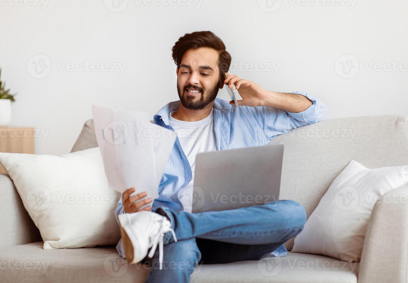 Eastern Freelancer Man With Cellphone, Papers And Laptop Working Remotely At Home. Millennial Arab Guy Reading Documents And Talking On Mobile Phone While Sitting On Couch With Computer, Free Space photo