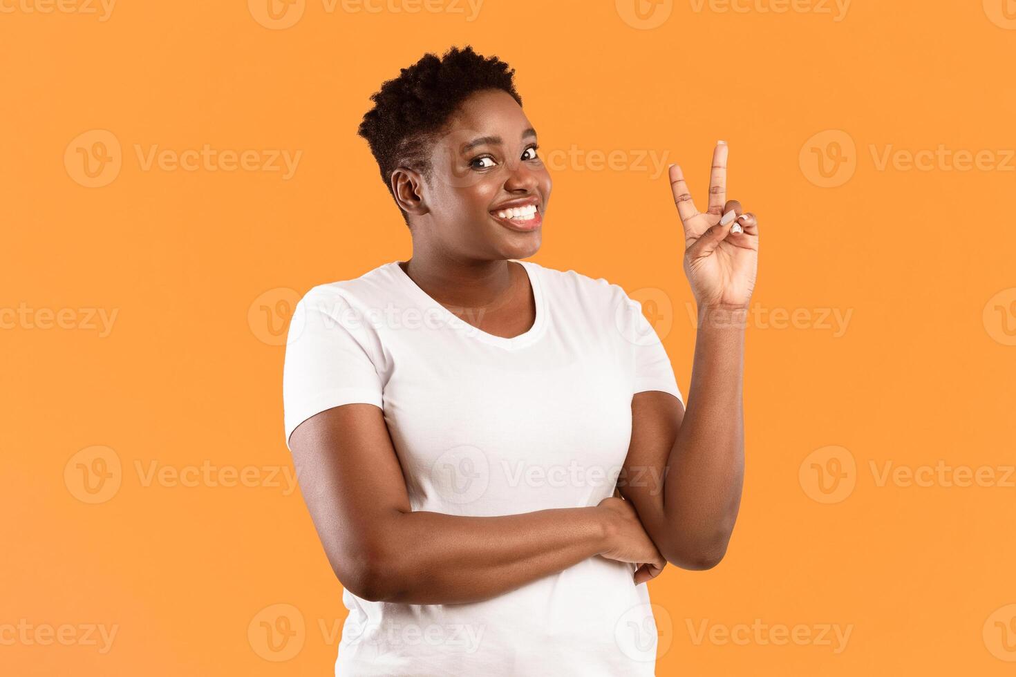 Smiling Oversized Black Woman Gesturing V-Sign To Camera Standing Over Yellow Background. African Female Showing Victory Gesture Smiling Posing In Studio photo