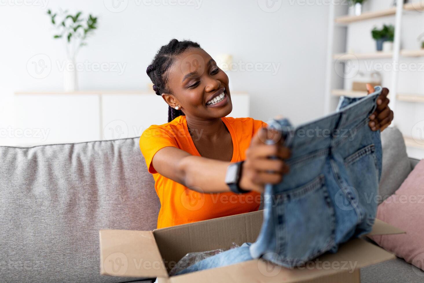 Ordering clothes online. Smiling black female customer opening parcel box, looking at new jeans on sofa at home. Young Afro woman satisfied with internet shopping and delivery service photo