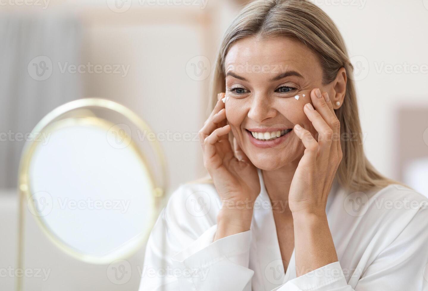 Happy beautiful blonde middle aged lady in silky bathrobe using moisturizing eye cream, looking at mirror and smiling, enjoying face care routine, closeup shot. Dark circles, eye wrinkles treatment photo