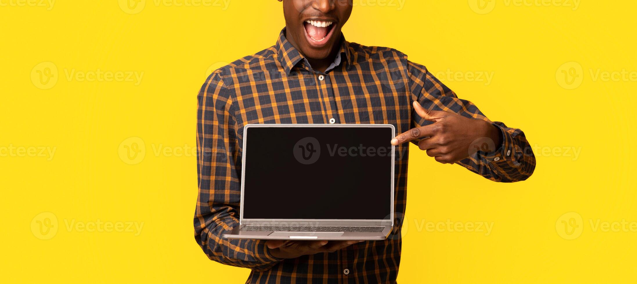 Check This Website. Joyful African Guy Pointing At Laptop Computer With Black Blank Screen, Standing Isolated Over Yellow Background, Demonstrating Free Place For Your Design, Mockup With Copy Space photo