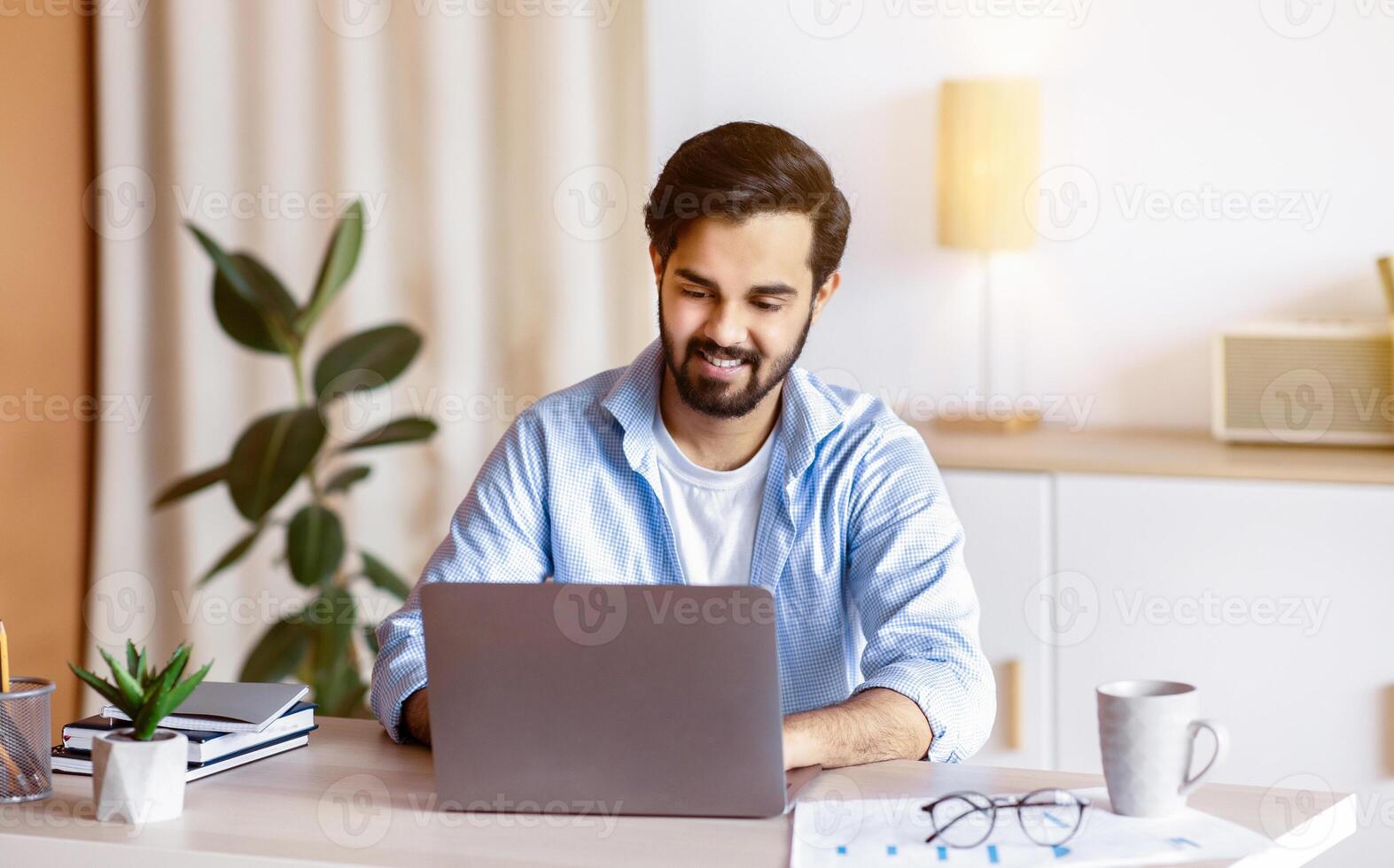 Cheerful Middle Eastern Man Sitting At Desk In Home Office And Working On Laptop. Handsome Arab Male Entrepreneur Using Computer, Enjoying Remote Distance Job Opportunities, Free Space photo