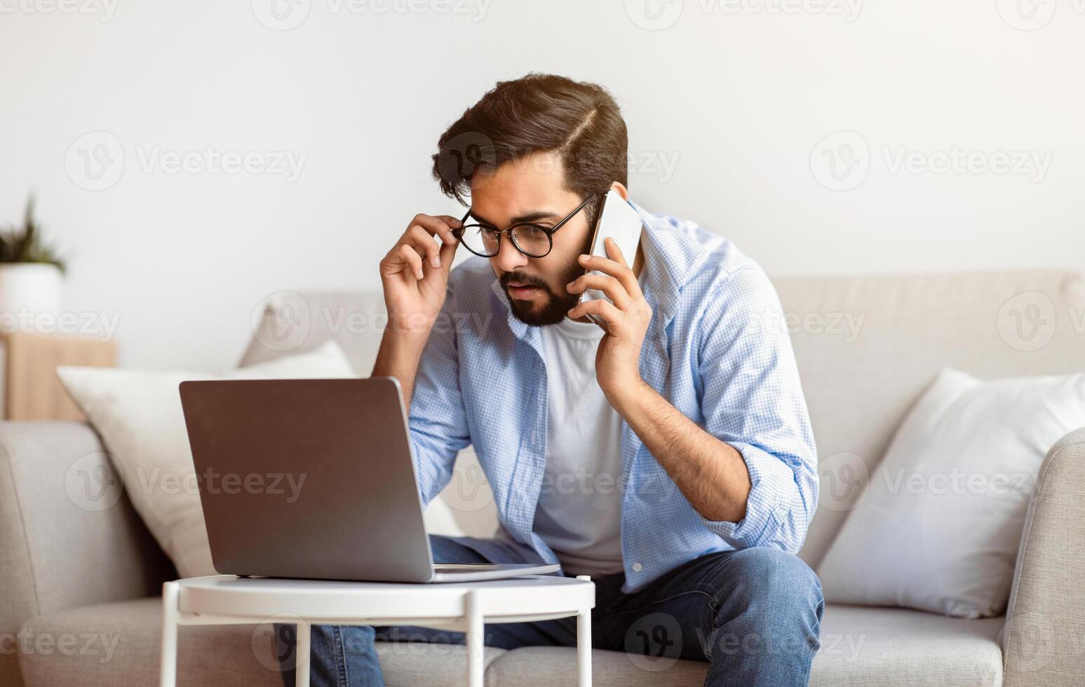 Arab Freelancer Man Talking On Cellphone And Working With Laptop At Home Office, Busy Eastern Guy Looking At Computer Screen While Discussing Project With Client, Sitting On Couch In Living Room photo