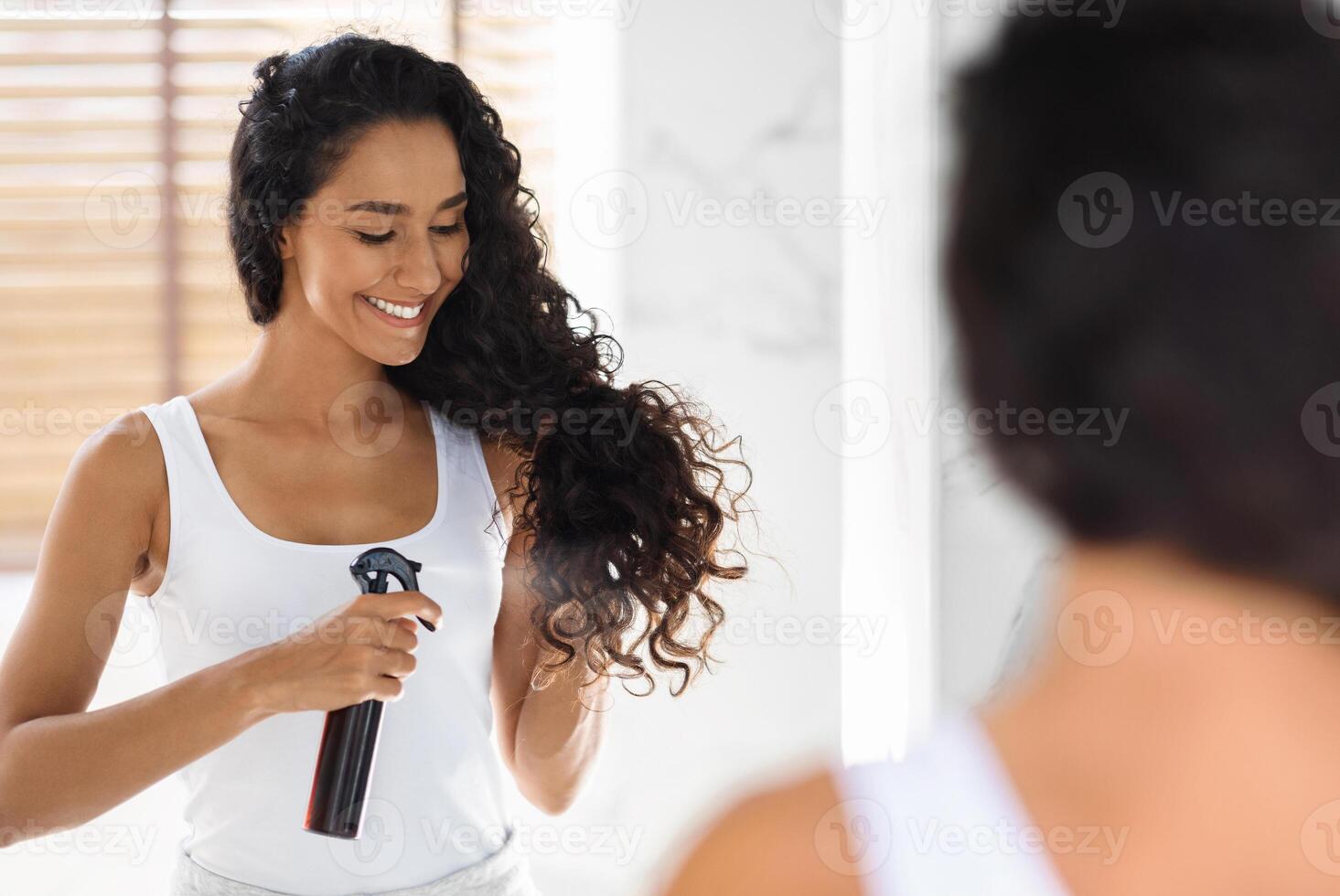 Haircare. Beautiful Young Lady Applying Hair Spray For Curls While Standing Near Mirror In Bathroom, Smiling Millennial Woman Using New Hairstyle Product At Home, Selective Focus On Reflection photo