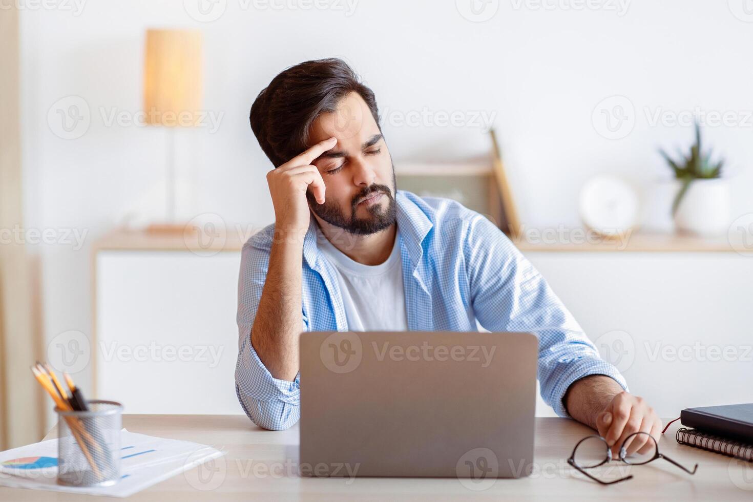 Overworked Arab Businessman Suffering From Migraine At Work, Having Strong Headache. Stressed Millennial Eastern Man Feeling Unwell While Sitting At Workplace In Modern Office, Copy Space photo