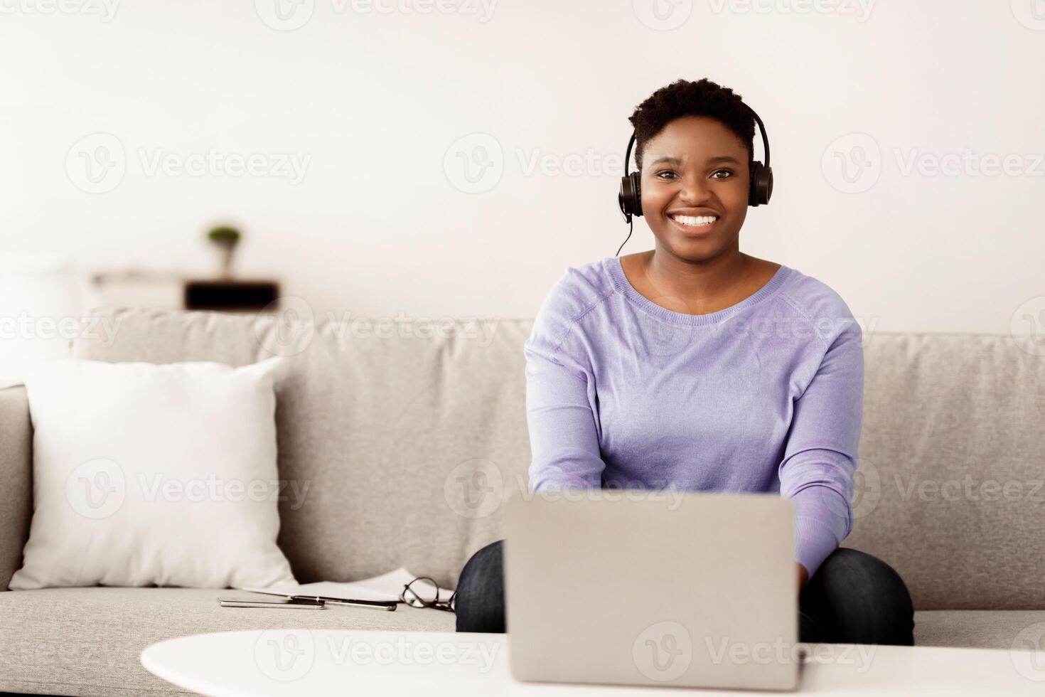 Portrait of smiling female customer service operator in headset posing at workplace, looking at camera, portrait of body positive hotline manager sitting on sofa with laptop in home office, copy space photo