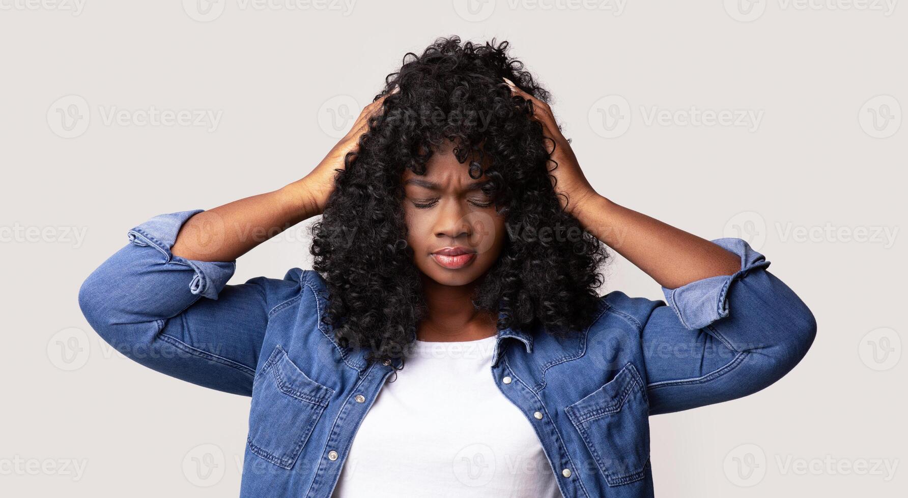 Upset young black lady in jeans coat and white t-shirt with curly hair having headache, touching her head, closing eyes, grey studio background. Migraine, headache, tension, cluster concept photo
