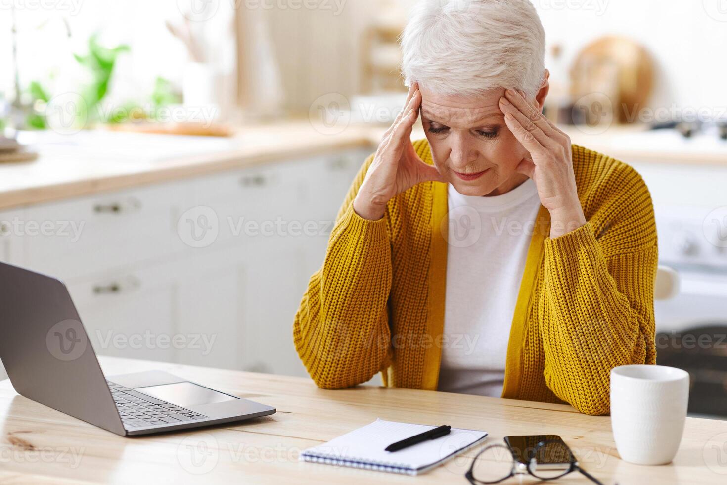 Tired mature grey-haired woman freelancer working from home, using laptop, sitting in kitchen, copy space. Stressful senior lady taking notes, sitting in front of computer, touching her head photo