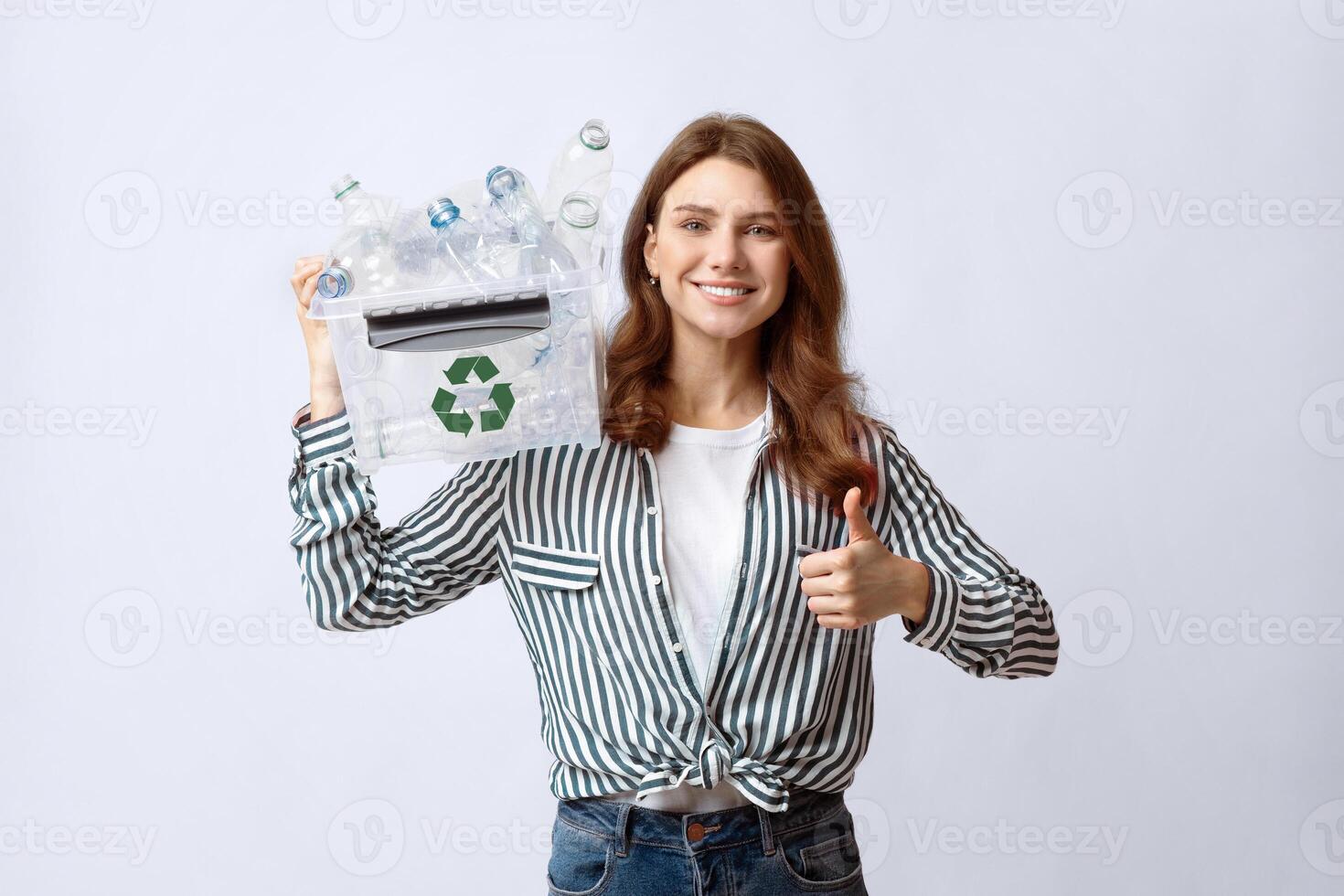Waste Recycling Concept. Smiling young woman holding recycle bin with empty plastic bottles and showing thumb up gesture, eco-friendly lady standing over light background in studio, copy space photo