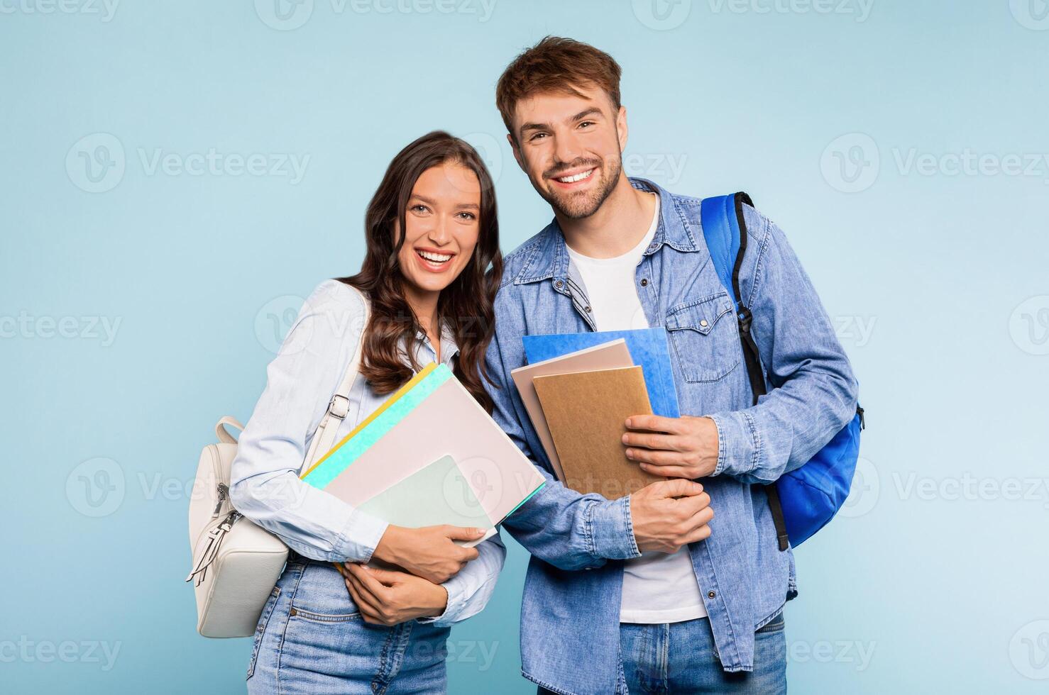 Happy young student couple in denim, with man wearing backpack and holding books, and woman clutching folders, both ready for day at university, against bright blue background photo
