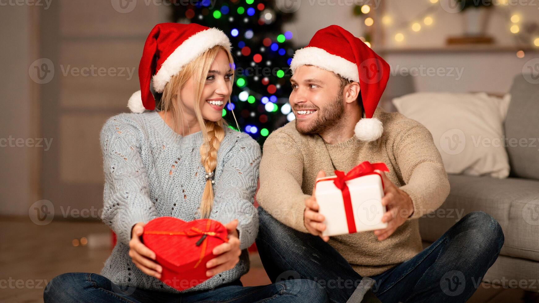 Loving family husband and wife celebrating New Year together at home, holding presents, looking at each other and smiling, empty space. Happy man and woman in Santa hats with gift boxes photo