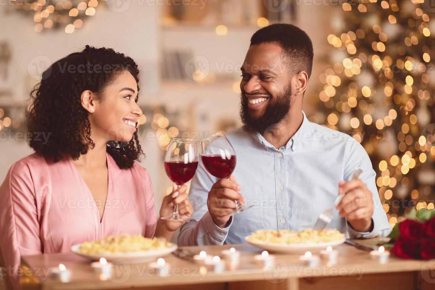 Celebrating St Valentine's Day. Happy loving african american man and woman sitting at table in restaurant or at home, drinking red wine, holding glasses, having romantic dinner, looking at each other photo
