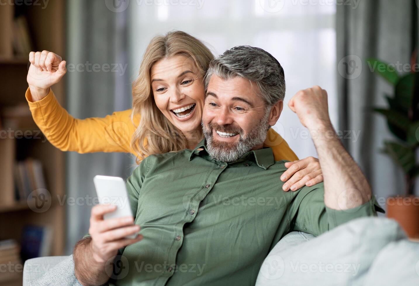 Excited Middle Aged Couple Celebrating Success While Looking At Smartphone Screen At Home, Joyful Mature Spouses Cheering Win, Emotionally Reacting To Good News, Raising Fists With Excitement photo